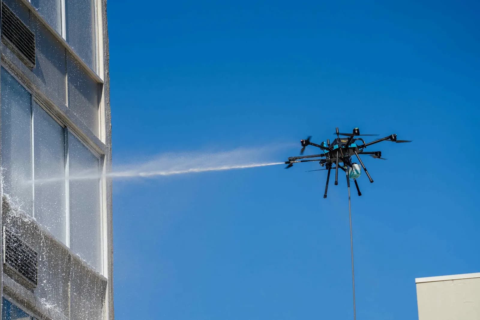 Tethered cleaning drone spraying water across the windows of a high-rise building against a clear blue sky