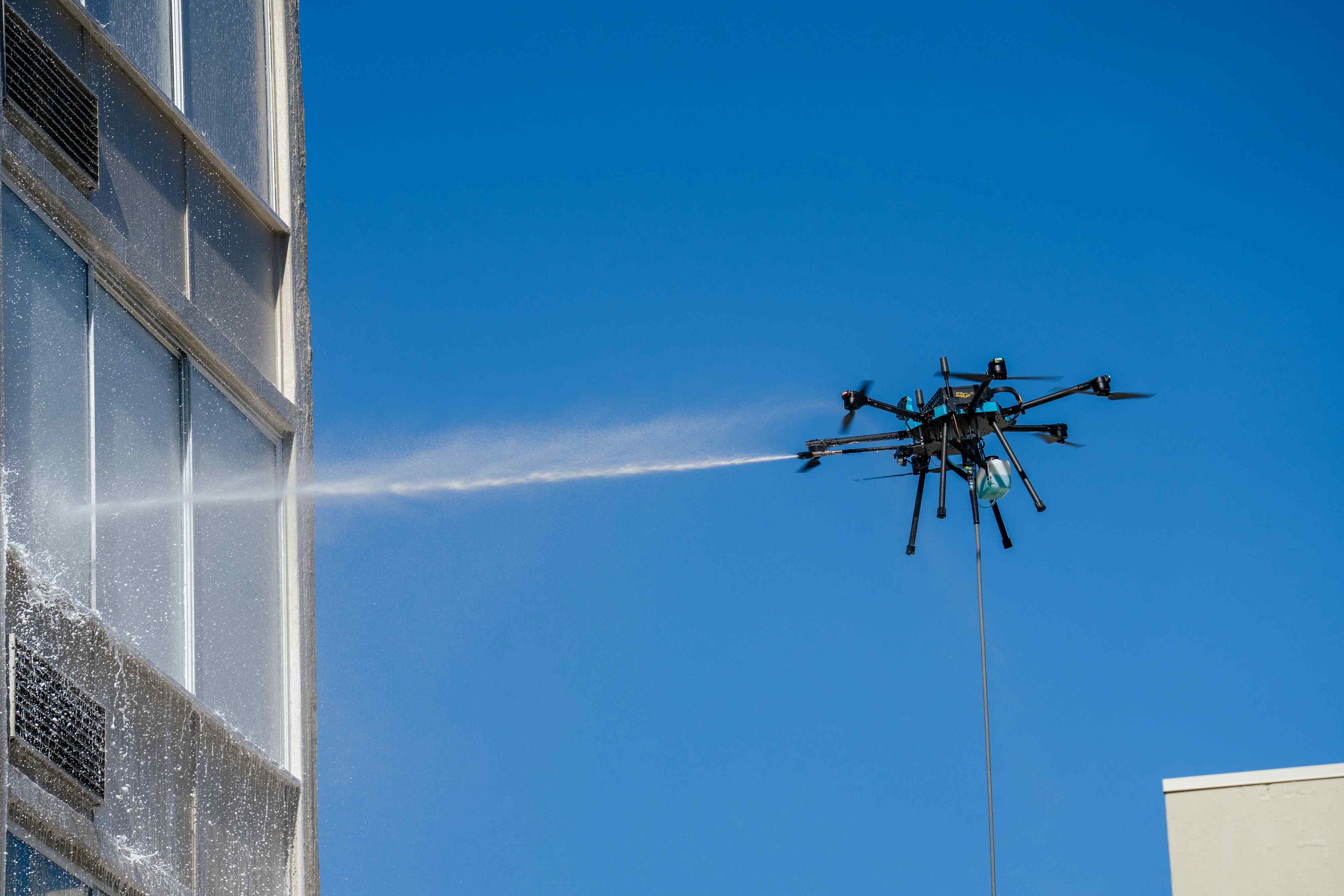 Tethered cleaning drone spraying water across the windows of a high-rise building against a clear blue sky